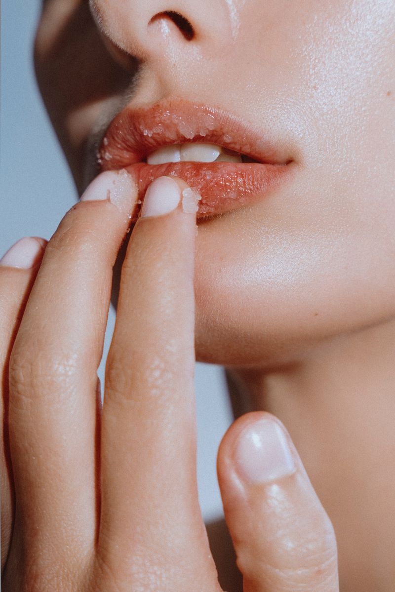 Close-up shot of an adult woman's face featuring fair skin, light pink lips, and fingers touching lips against a soft blue background.