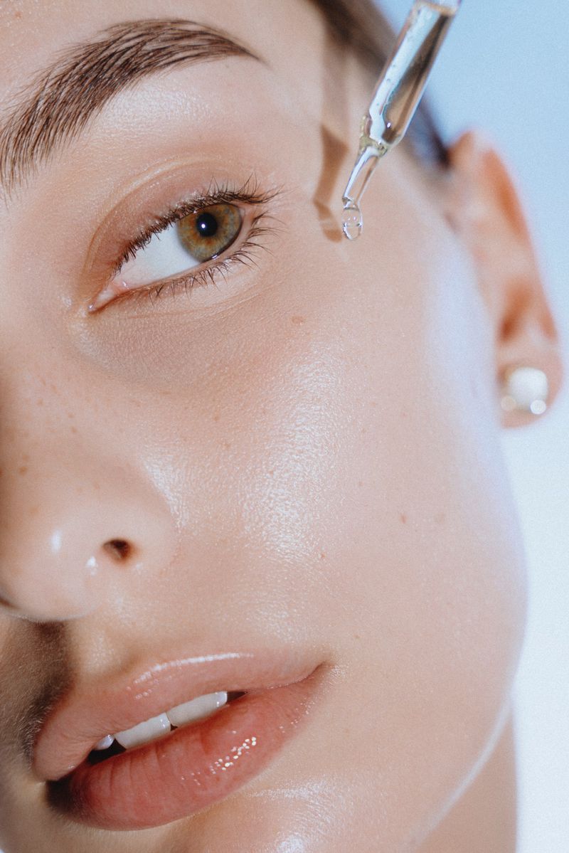 Close-up of a fair-skinned woman receiving a clear serum application from a dropper onto her face in front of a light blue background.