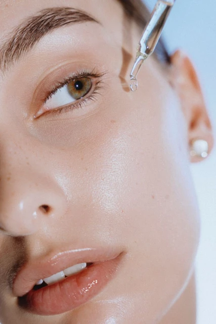 Close-up of a fair-skinned woman receiving a clear serum application from a dropper onto her face in front of a light blue background.