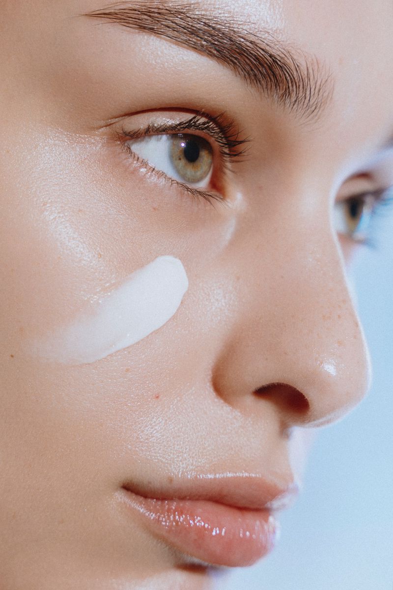 Close-up of a woman's face with a streak of white cream on her cheek in a beauty shot on a light blue background.