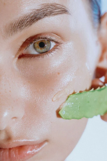 Close-up of a woman's face with a slice of aloe vera leaf applying gel on her cheek. Focus on skincare and natural beauty.