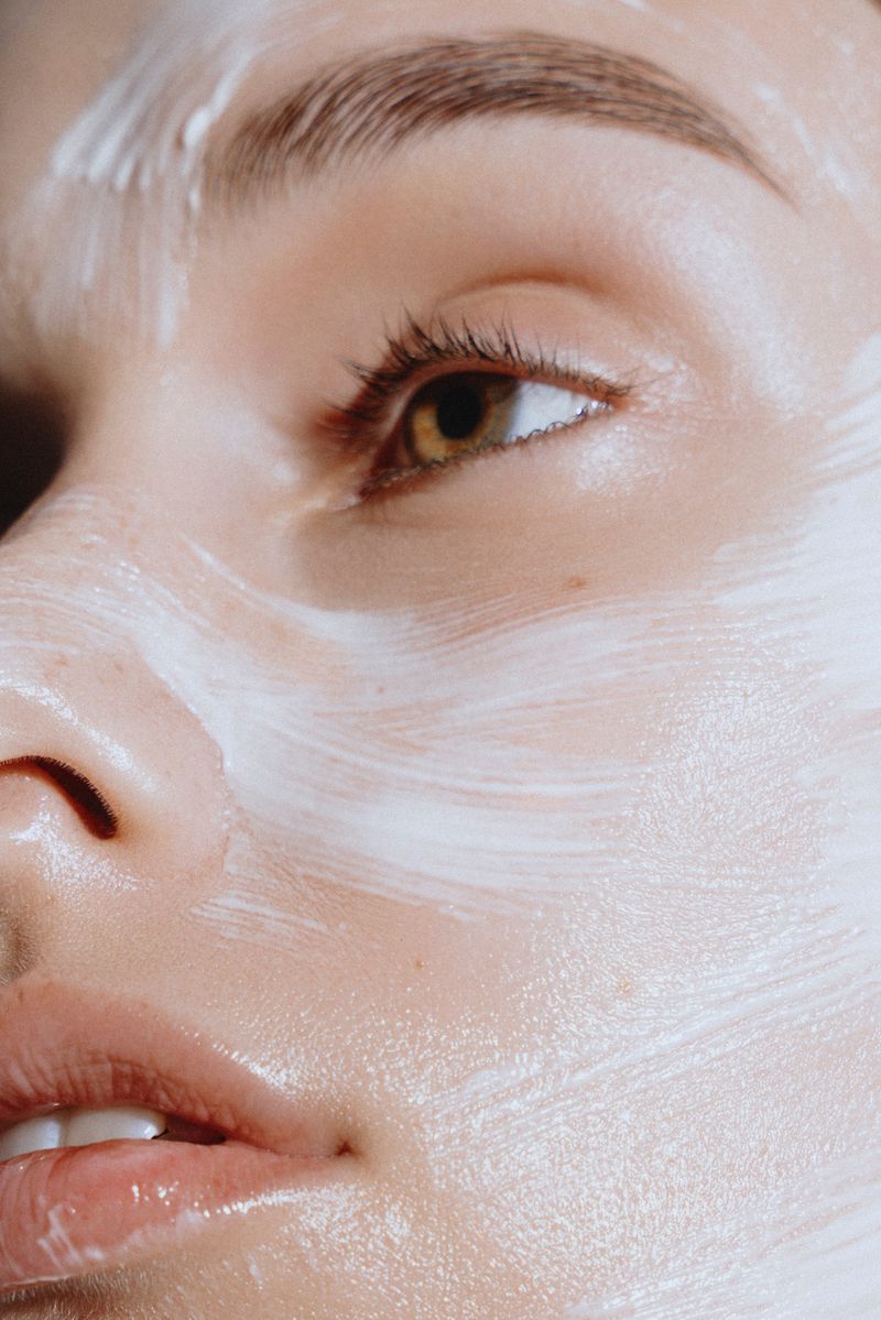 Close-up of a woman's face with white skincare cream applied in textured brush strokes across her cheek, nose, and forehead.