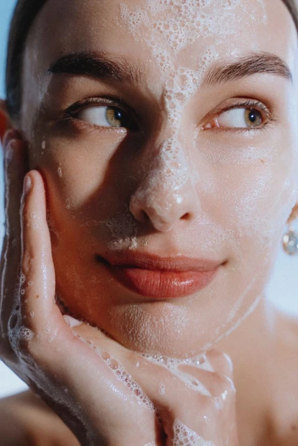 Close-up of a woman with soap on her face and hand, looking away, in a beauty or skincare context with soft, diffused lighting.