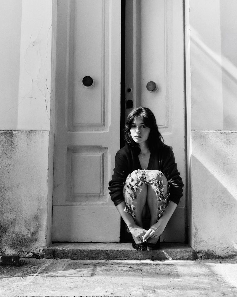 A young woman with dark hair is crouching in front of a white door in a monochromatic outdoor shot with a soft lighting and neutral tones.