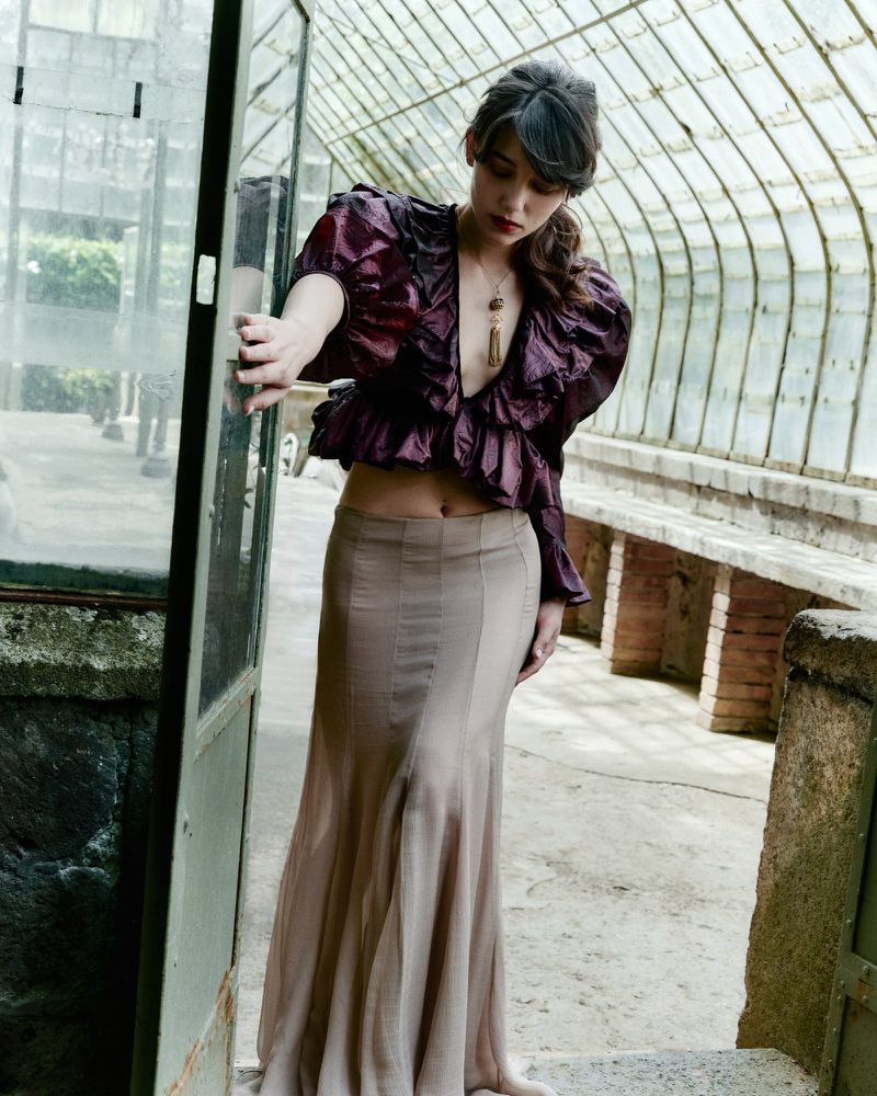 Woman poses in greenhouse wearing ruffled top and flowing skirt. Natural light illuminates glass structure, stone details, and verdant plants.