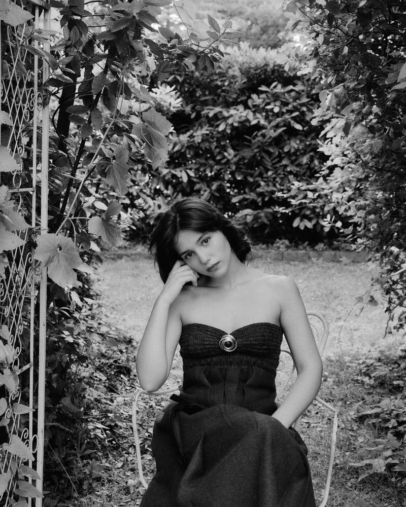 Monochrome portrait of a young woman with dark hair in a strapless dress, seated in a garden with lush greenery and a decorative metal gate.