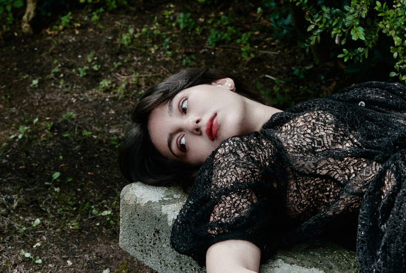 Young woman with dark hair and red lipstick lies on a stone surface in a black lacy top, looking away in an outdoor setting.