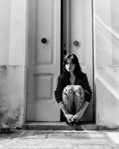 Black and white portrait of a woman crouching in a tall doorway holding a cigarette while looking directly at the camera.