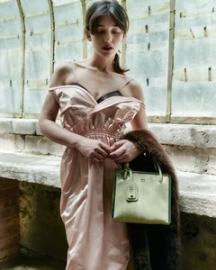 Woman in a draped blush pink satin dress holding a sage green leather handbag and brown fur wrap against a weathered glass window.