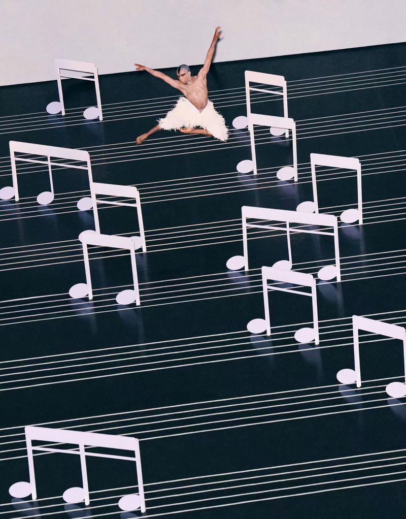 Male dancer in white costume leaps on a stage with music note props and black and white stripes mimicking sheet music.