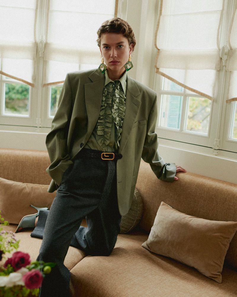 An adult female with short brown hair poses indoors wearing a green blazer and frilled blouse with a tan couch and natural window light.