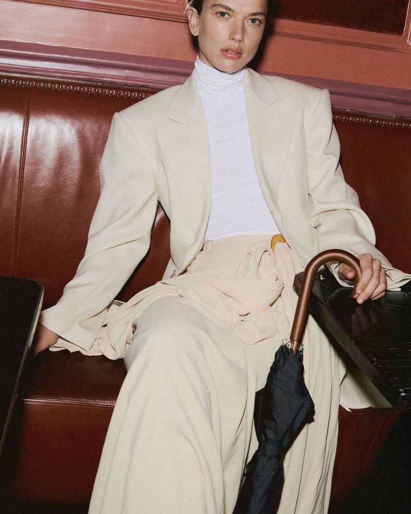 Adult posing indoors while seated. Dressed in a white turtleneck, blazer, pants, and holding a closed black umbrella.