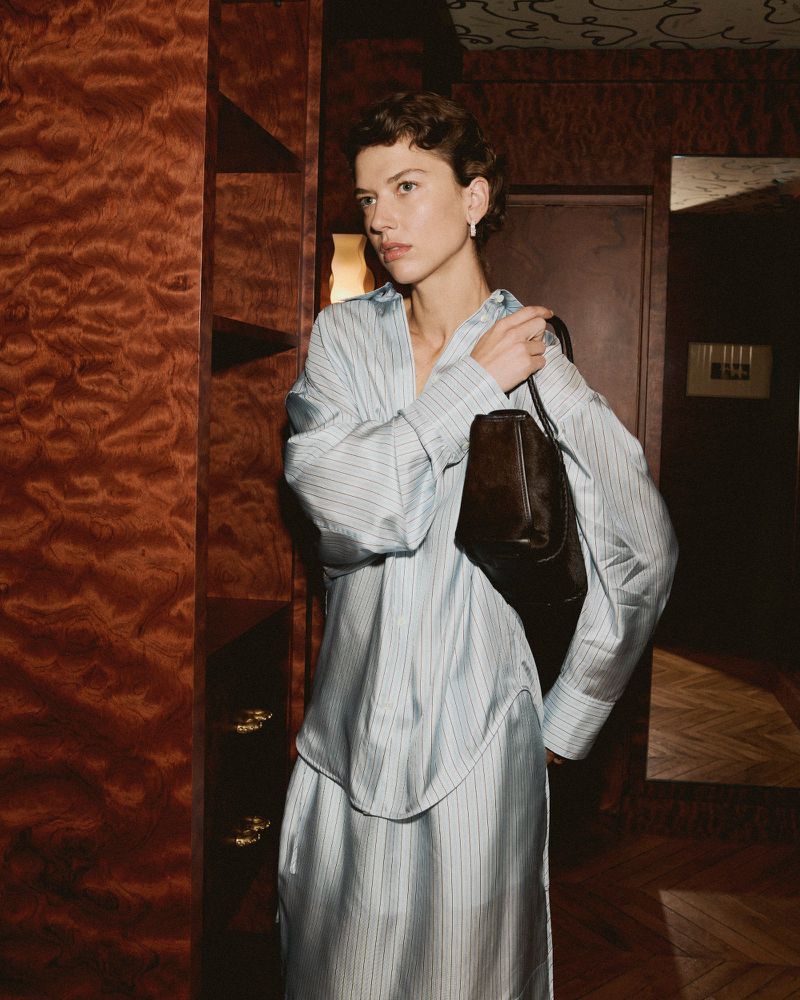 Woman in blue striped dress with brown leather bag poses indoors against wood paneled wall, showcasing fashion and style.