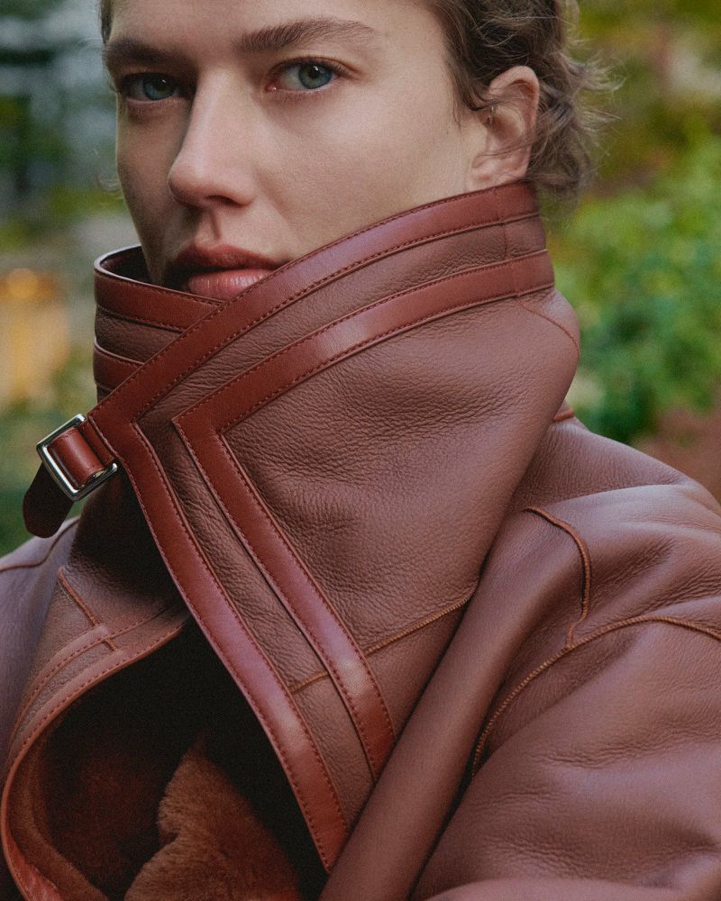 Close-up portrait of a fair-skinned woman in a brown leather shearling coat, set against a blurred background of trees and foliage.