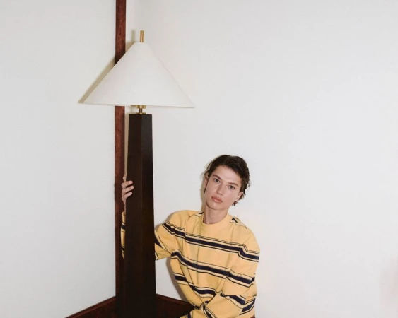 Young adult in a yellow striped long-sleeve shirt poses in an indoor corner setting, with a floor lamp behind. Simple and clean background.