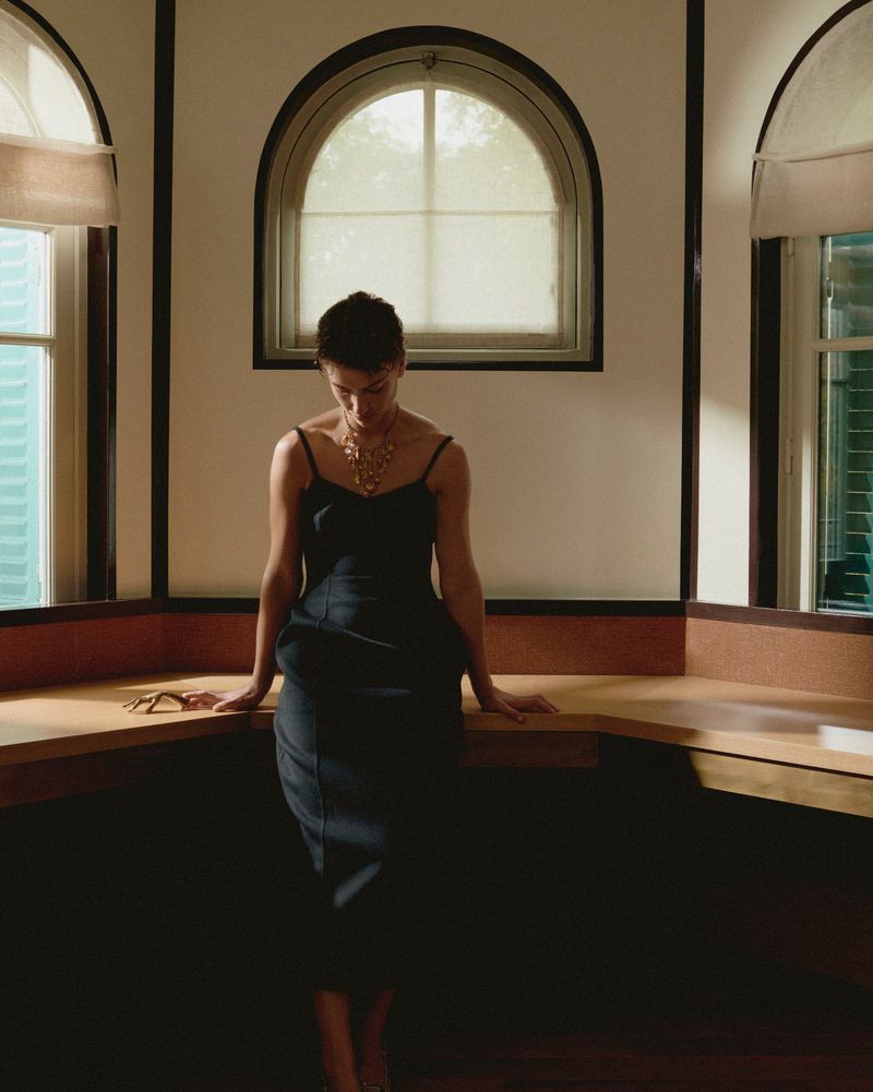 A woman in a black dress and gold necklace stands inside near arched windows, casting shadows on her and the counter behind her.