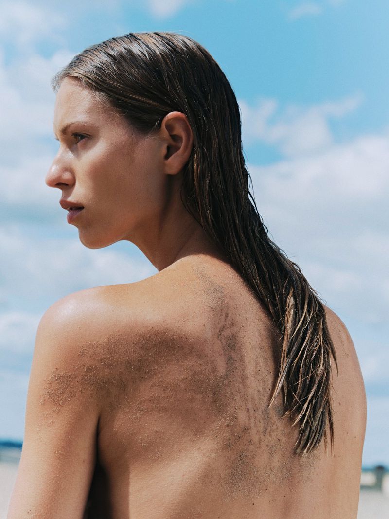 Model Lea Seelenmeyer with slicked wet hair and sand on her bare back viewed in profile against a bright blue sky.