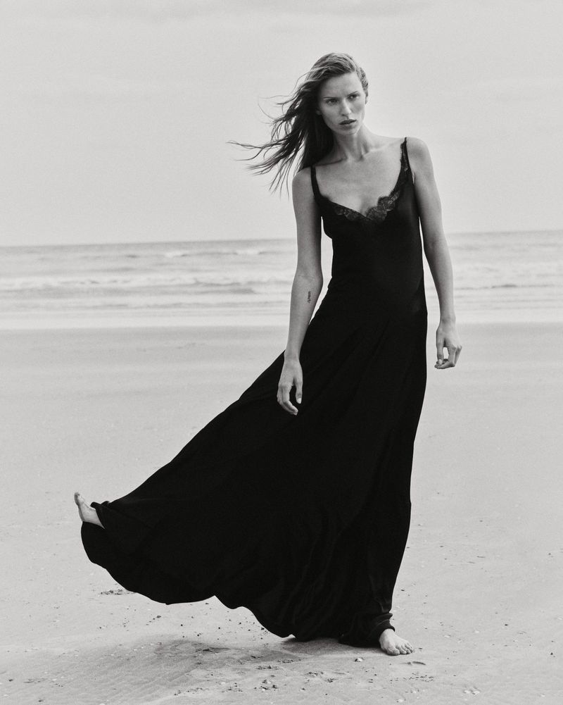 Model Lea Seelenmeyer standing barefoot on a sandy beach wearing a long black slip dress in a monochrome editorial photograph.