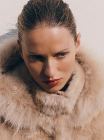 Lea Seelenmeyer wearing a plush beige shearling coat with a voluminous collar, looking downward against a pale textured background.
