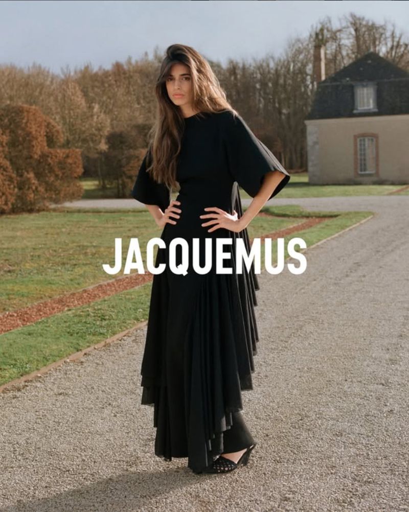 Brunette woman in a black dress stands with hands on hips, posing on a gravel path in front of trees and a building with the brand name.