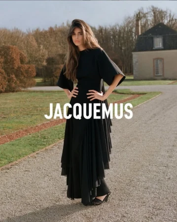 Brunette woman in a black dress stands with hands on hips, posing on a gravel path in front of trees and a building with the brand name.