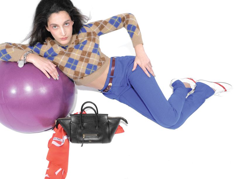 Woman in blue pants and patterned sweater poses with a purple exercise ball and black bag in a studio, fashion photography.