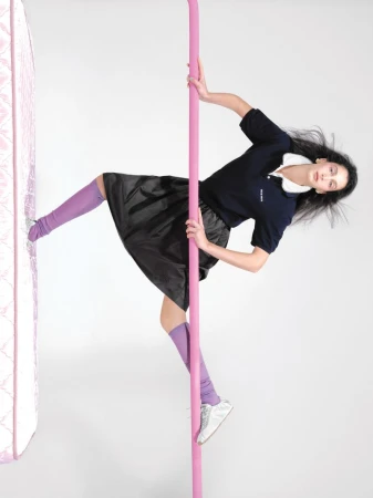 Stylish woman poses with a pink pole in a studio. She wears a navy polo shirt, black skirt, purple socks, and silver sneakers on a white background.