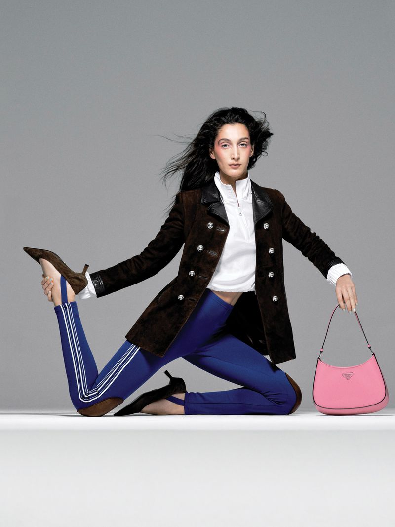 A fashion shot of a woman with long dark hair wearing a brown blazer and blue leggings holding a pink Prada handbag in a studio.