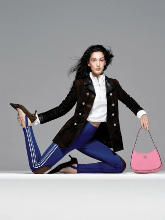 A fashion shot of a woman with long dark hair wearing a brown blazer and blue leggings holding a pink Prada handbag in a studio.