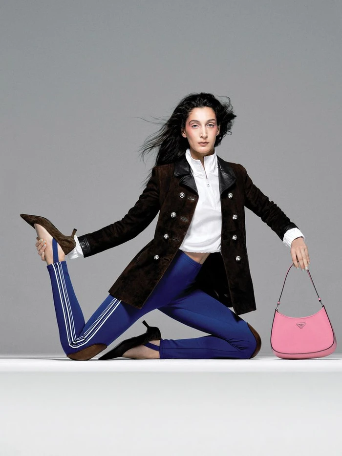 Model Maya Mani poses dynamically in a dark brown buttoned coat and blue stirrup leggings while holding a pink handbag in a studio.