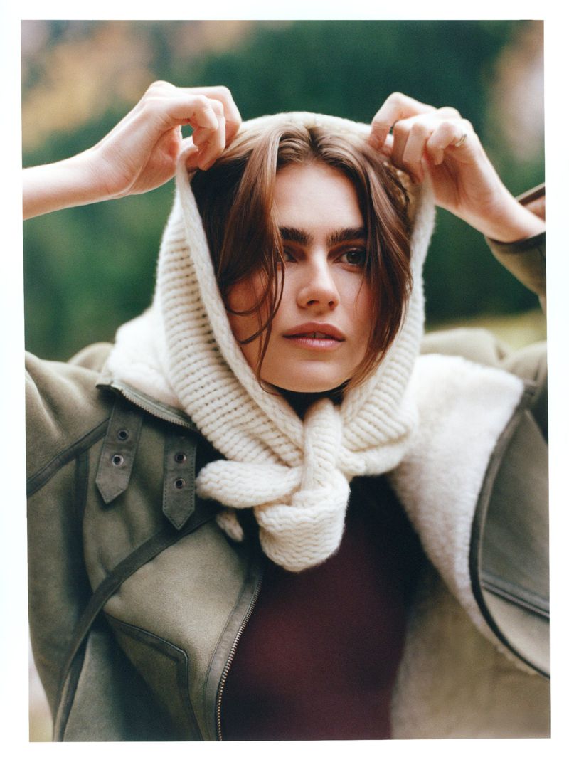 Model Mia Armstrong wearing a white knit hood scarf and olive green shearling jacket with hands raised to her head outdoors.