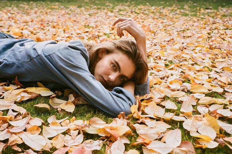 Young woman lies on a bed of colorful fallen leaves on a sunny day in autumn, looking directly at the camera with a relaxed expression.