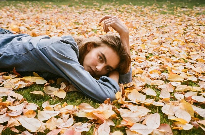 Young woman lies on a bed of colorful fallen leaves on a sunny day in autumn, looking directly at the camera with a relaxed expression.