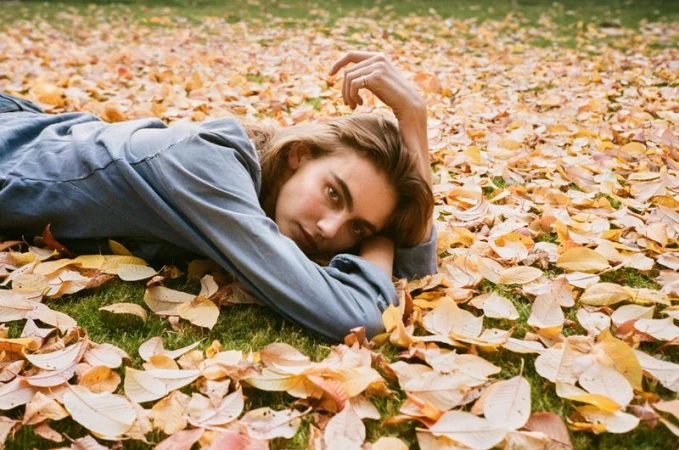 Young woman lies on a bed of colorful fallen leaves on a sunny day in autumn, looking directly at the camera with a relaxed expression.