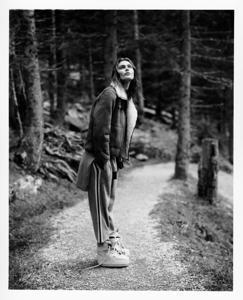 A black and white portrait shows a woman in a jacket and sweatpants looking up while standing on a path lined with tall trees.