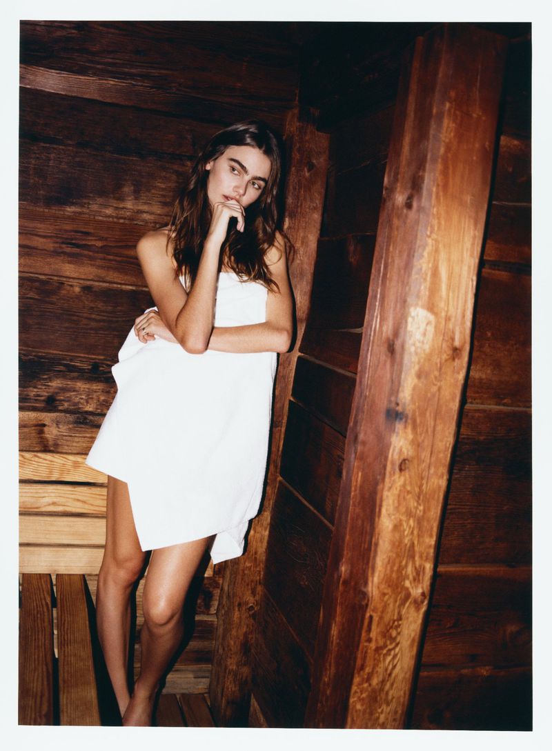 Young woman draped in a white towel poses against a wooden sauna wall, looking at the camera in soft, natural light.