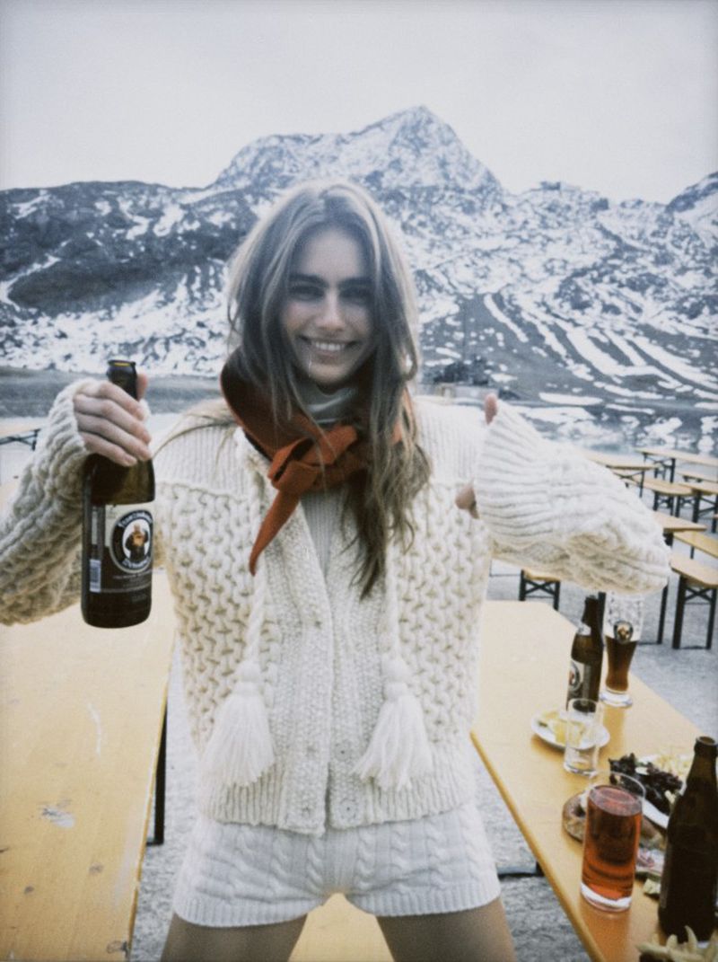 Smiling woman poses outdoors with a beer bottle, wearing a knitted sweater and shorts, with snow-covered mountains in the background.