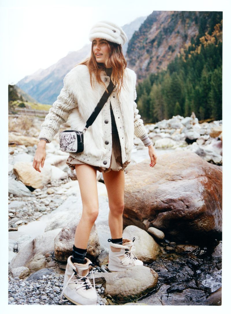 A young woman poses on rocks near a stream, wearing a cream-colored hat, jacket, and boots with a patterned shoulder bag, set against a mountain backdrop.