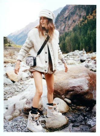 A young woman poses on rocks near a stream, wearing a cream-colored hat, jacket, and boots with a patterned shoulder bag, set against a mountain backdrop.