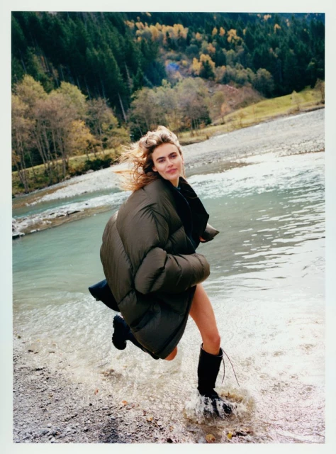A blonde woman in a puffy coat runs through a shallow river with mountains and trees in the background on an overcast day.