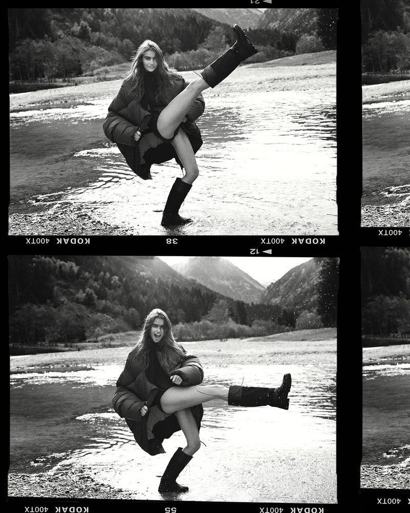 Black and white shot of a woman in a puffy jacket and boots in a shallow body of water with mountains in the background.