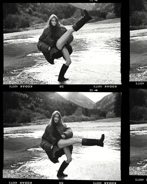 Black and white shot of a woman in a puffy jacket and boots in a shallow body of water with mountains in the background.