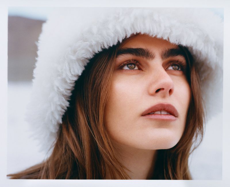 Portrait of a woman with light brown hair, wearing a white fur hat, and looking up towards light outdoor background. Close-up shot.
