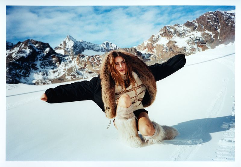 Female model wearing a fur-lined hooded jacket and furry boots poses in a snowy mountain landscape under a clear blue sky.
