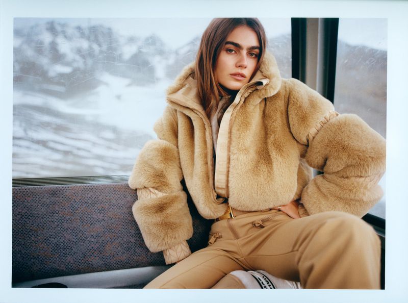 A female model poses indoors wearing a furry beige jacket and beige trousers. The background shows a snowy mountain scene.
