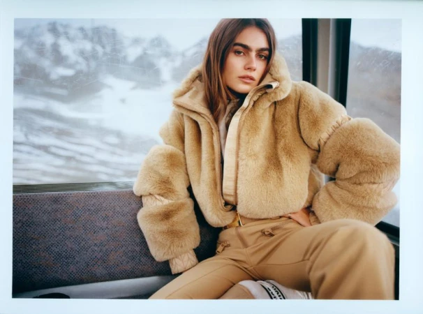 A female model poses indoors wearing a furry beige jacket and beige trousers. The background shows a snowy mountain scene.