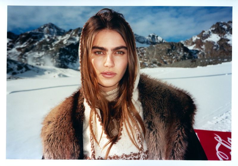 Close-up portrait of a young adult woman with light brown hair and fur-trimmed coat, against a snowy mountain backdrop on a sunny day.