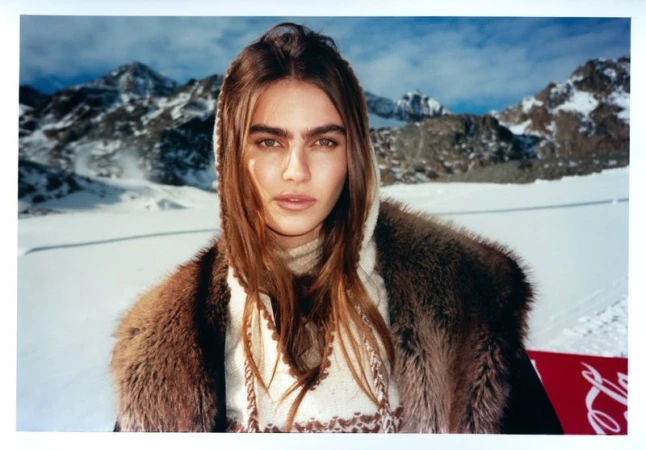Close-up portrait of a young adult woman with light brown hair and fur-trimmed coat, against a snowy mountain backdrop on a sunny day.