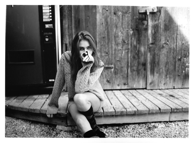 Monochrome portrait of a young woman in sweater sitting on wooden porch with weathered wooden wall and vending machine background.