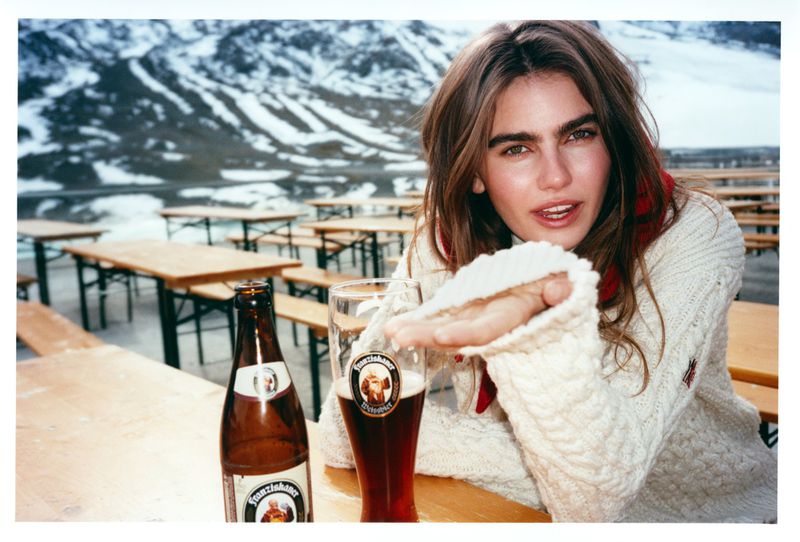 Blonde woman in a cable knit sweater sits at a wooden table with two beers. Mountains are blurred in the background.